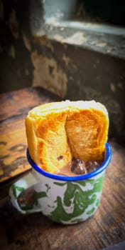 Ice chocolate drink on a vintage Indonesian iron mug with random green pattern called Blirik cup or cangkir Blirik on bokeh background along with chocolate filling bread on a cafe in Bogor