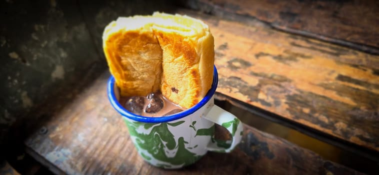Ice chocolate drink on a vintage Indonesian iron mug with random green pattern called Blirik cup or cangkir Blirik on bokeh background along with chocolate filling bread on a cafe in Bogor