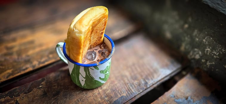 Ice chocolate drink on a vintage Indonesian iron mug with random green pattern called Blirik cup or cangkir Blirik on bokeh background along with chocolate filling bread on a cafe in Bogor