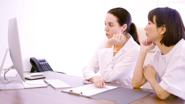 Two female doctor talking about medical results while using computer sitting on a desk in a clinic