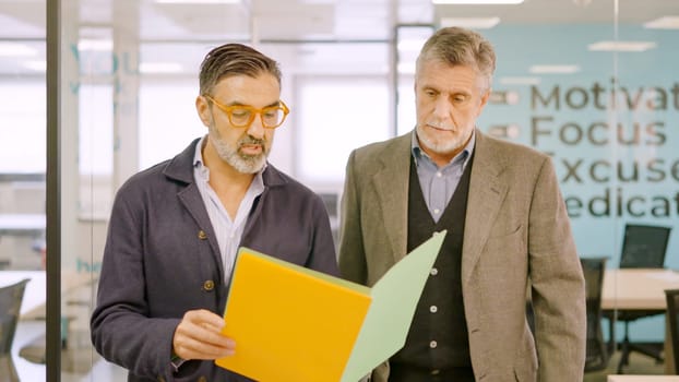 Two mature businessmen walking while reading a financial report on paper in a coworking space
