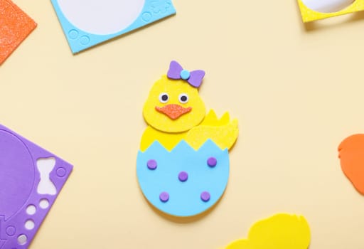 Colorful felt easter chick made by child's hands lies on a light yellow background with used stickers on the children's table, flat lay closeup. The concept of crafts, diy, needlework, diy, children art, artisanal, Easter preparation,children creative.