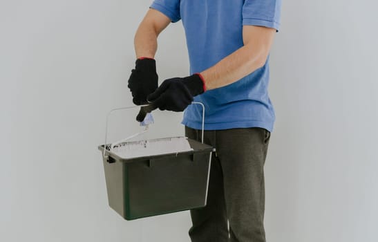 One young unrecognizable Caucasian man in a blue T-shirt and gray pants holds a black bucket with white paint and dips a roller in it, standing against a white wall, close-up side view.
