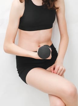 A young caucasian teenager girl holds a black dumbbell with one hand resting her back against a white wall, close-up side view. The concept of home fitness and healthy lifestyle.