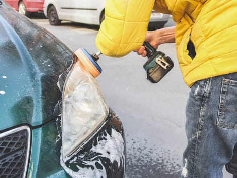 A young caucasian guy without a face in a yellow jacket is being drilled with a sponge disc and polishing the headlight of his car with a detergent soap on the city street in front of the house, close-up side view.Concept home car wash and cleaning with polishing car headlights.