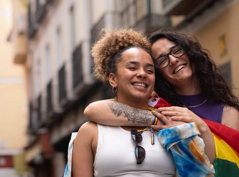 Two multiethnic happy lesbian women are hugging and smiling enjoying time in Madrid city street.