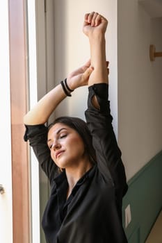 Young model, dressed in a black shirt, stretching her arms over her head by the window, with her eyes closed.