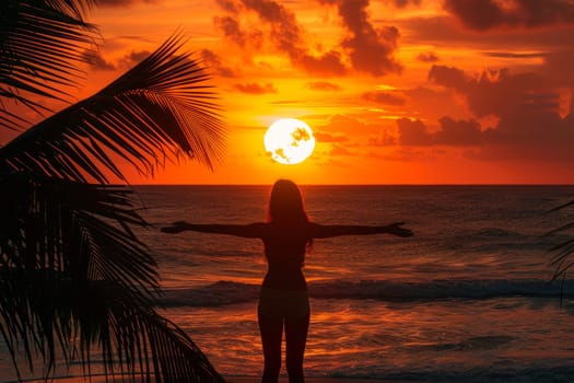 Silhouette of woman with arms outstretched enjoying tropical sunset beach with palm trees and dramatic orange sky. Concept of relaxation, freedom, and paradise