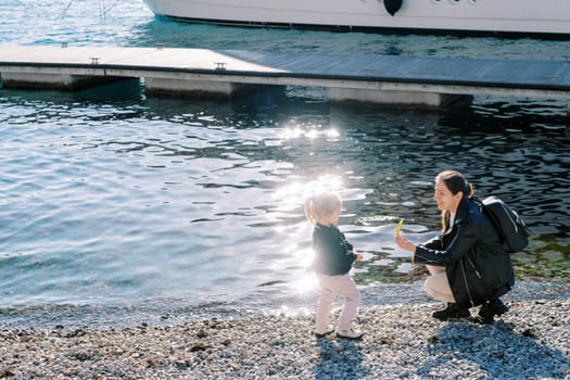 Mom hands a green leaf to a little girl squatting on a pebble beach. High quality photo