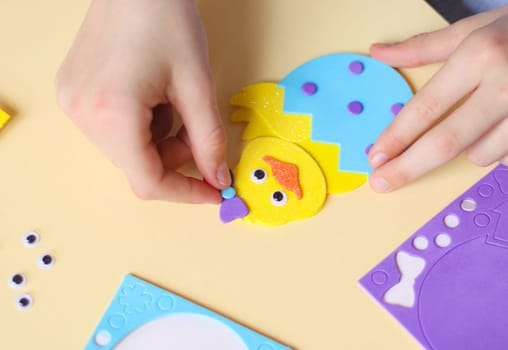 The hands of a caucasian teenage girl stick a lilac bow sticker on a yellow chicken felt with her fingers, sitting at a children's table with a set of handmade items on a pale yellow background with selective focus, flat lay closeup. The concept of crafts, diy, needlework, diy, children art, artisanal, Easter preparation,children creative.