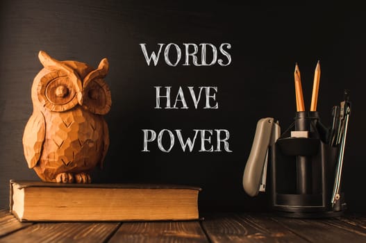 A wooden owl sits on top of a stack of books. The owl is surrounded by writing that says Words have power. The image conveys the idea that words can have a significant impact on people