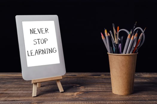 Never stop learning is written on a white sign. A wooden stand holds a white tablet and a cup of pencils