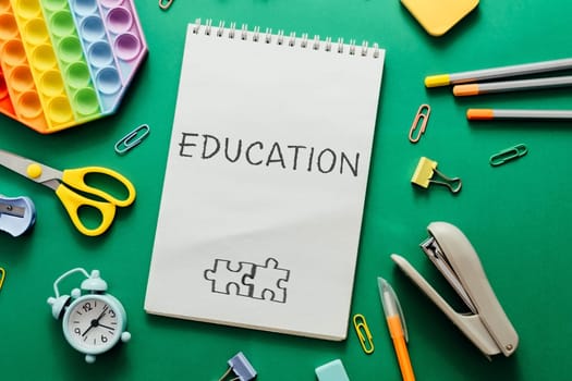 A notebook with the word education written on it. The notebook is on a green surface with various school supplies such as scissors, a stapler, and a clock