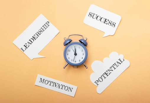 A blue alarm clock sits on a table with a bunch of paper cutouts around it. The paper cutouts spell out the words leadership, motivation, success, potential