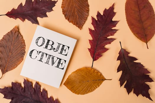 A white piece of paper with the word objective written on it. The paper is placed on top of a pile of autumn leaves, which are scattered around the paper. The leaves are of various sizes and colors