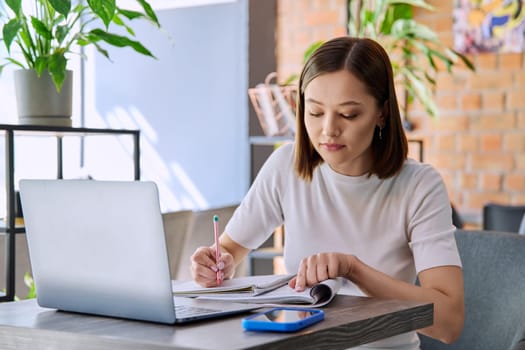 Young female college student studying using laptop, writing in notebook while sitting in coworking cafe. E-learning, education, lifestyle, youth concept