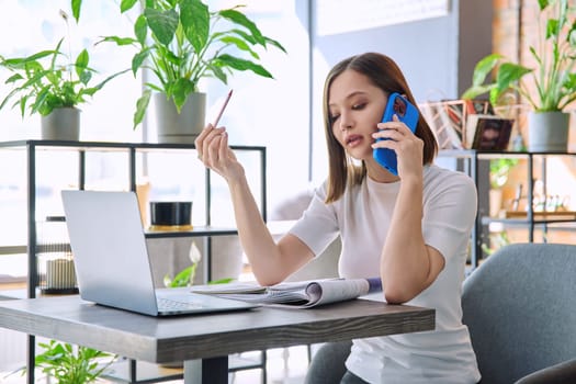 Young woman sitting at table with laptop computer talking on mobile phone in coworking cafe. Female university college student preparing for test exam, freelancer, business employee working remotely