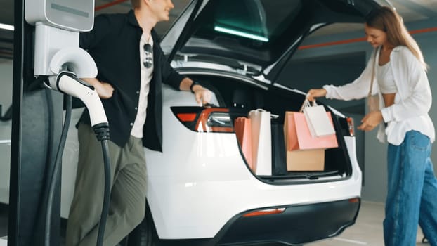 Young couple with shopping bag travel by EV electric car to shopping center parking lot charging in green city urban sustainability lifestyle by clean rechargeable energy of electric vehicle innards