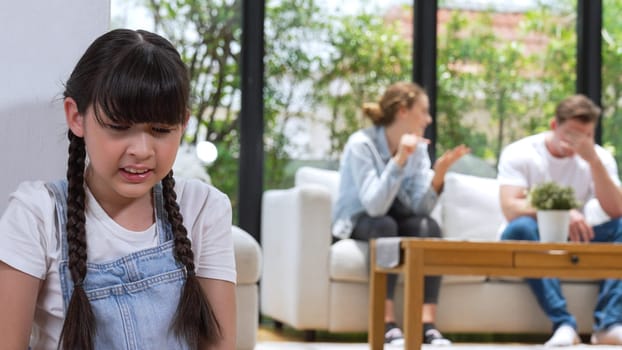 Stressed and unhappy young girl huddle in corner, cover her ears blocking sound of her parent arguing in background. Domestic violence at home and traumatic childhood develop to depression. Synchronos