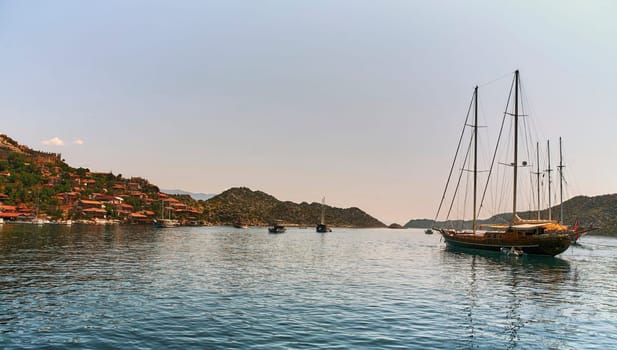 Yachts docked in a serene bay surrounded by lush green mountains. Clear water reflects the sun. Various yachts with people on board. Trees cover mountains, small town visible.