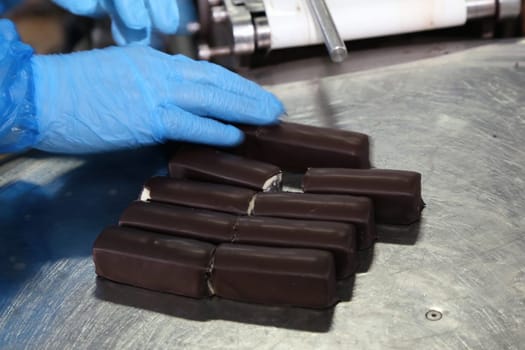 A gloved hand inspects a batch of freshly made chocolate bars on a stainless steel surface at a confectionery production facility. Each bar is aligned uniformly, awaiting the next phase of packaging or additional quality checks to ensure consistency and excellence.