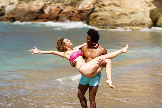 Side view of cheerful young female stretching arm backwards, while lying in hands of standing African American male and looking at each other near seawater during daytime