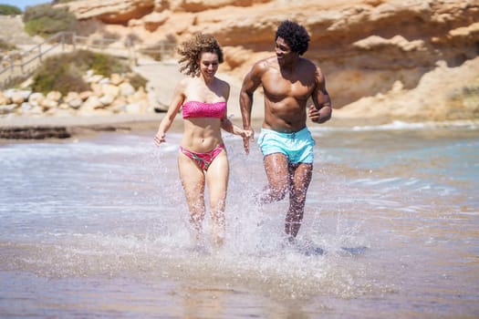 Full body of multiracial couple in swimwear running in splashing water and having fun while looking away and enjoying free time near seaside in summertime