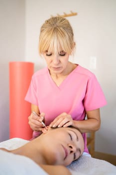 Chiropractor professional beautician doing deep ear massage of young female customer with ear probe pen during skin care treatment in modern beauty salon