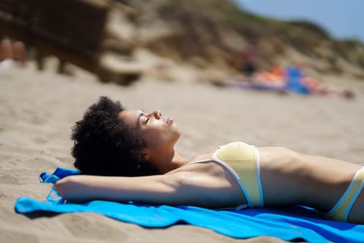 Side view of calm young African American female traveler with curly hair, in stylish swimwear enjoying, sunny summer day while lying on blue towel on sandy beach with closed eyes