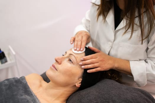 Crop cosmetician using cotton pad on face of female client for facial cleaning procedure in beauty salon during skin care treatment