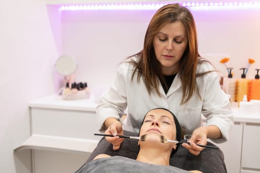 Beautician applying facial cleansing oil, with brush on woman lying face up with eyes closed during cosmetology session in beauty salon in background with cosmetics on counter