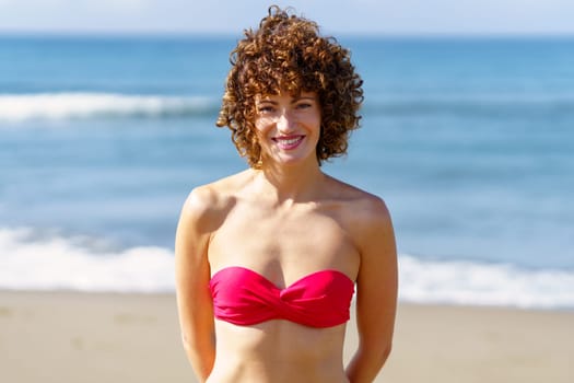 Portrait of happy young female in pink bikini with curly hair smiling and looking at camera while standing on beach near waving sea