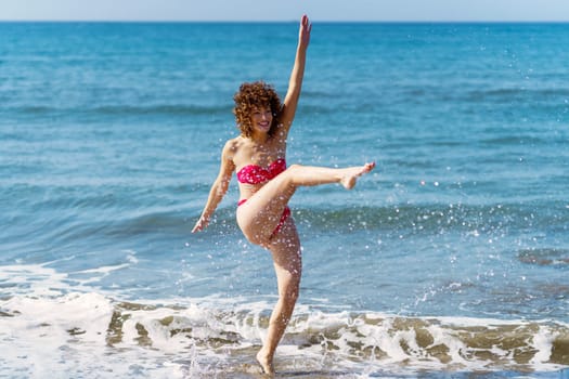 Full body of playful female in bikini kicking sea water and splashing on beach while enjoying sunny summer day