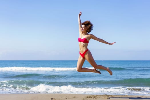 Full body side view of excited barefoot young female, in bikini with outstretched arms leaping with legs in air while looking away in daylight against rippling foamy seawater