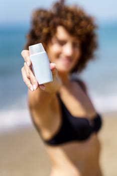 Soft focus of female in black bikini standing on beach near sea and demonstrating blank tube of sunscreen