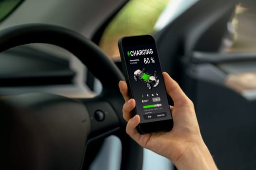 Young woman inside the vehicle, checking EV vehicle's application on battery recharging electricity status display on smartphone screen in modern EV car on her road trip journey. Exalt