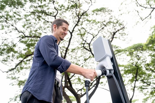 Man recharging battery for electric car during road trip travel EV car in natural forest or national park. Eco friendly travel during vacation and holiday. Exalt