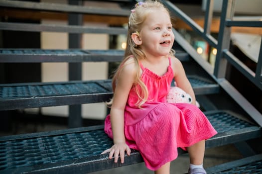 Child girl with hearing aid cochlear implant in pink dress having fun on summer street. Aid for the treatment of deafness and hearing loss in humans. Copy space.