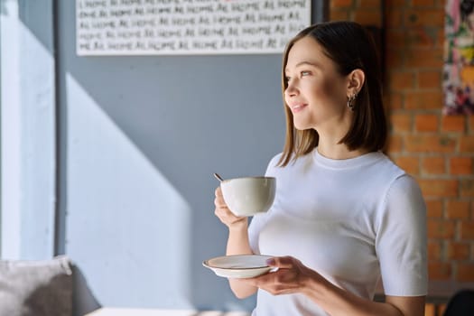 Young beautiful happy woman holding cup of coffee tea with saucer looking out window, gray wall of cafe coffee shop cafeteria copy space for text. Coffee business work services youth lifestyle leisure