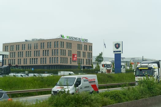 Seclin, France - May 22, 2023: A cluster of modern buildings with commercial signage, flanked by semi-trailers and vehicles on a busy highway during a clear day.