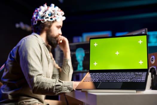 Man with EEG headset on writing code to transfer mind into virtual world, isolated screen laptop on desk Transhumanist using neuroscientific tech and chroma key notebook to gain superintelligence