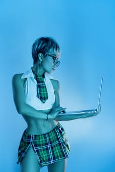 Sexy girl in glasses and an erotic school suit with a plaid skirt with a laptop in blue neon light on the background of copy paste