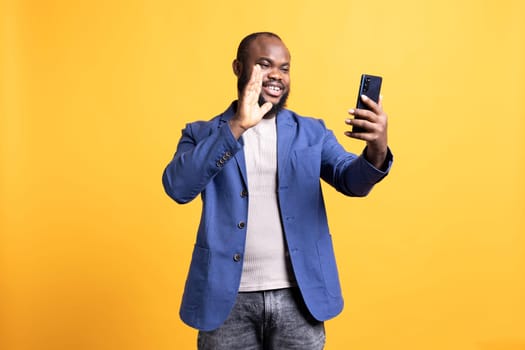 Joyful african american man greeting friends during teleconference meeting using smartphone, studio background. Happy person waving hand, saluting mate during internet video call