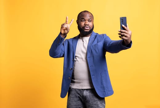 Man using smartphone to take selfies, doing victory hand sign. African american person taking photos using phone selfie camera, showing peace symbol gesturing, studio background