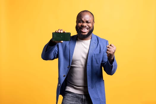 African american man excitedly celebrating while presenting isolated screen phone, studio background. Ecstatic BIPOC person holding copy space mockup cellphone used for advertising brands