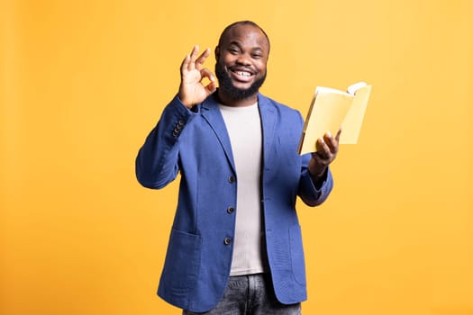 Portrait of smiling man recommending interesting book after being entertained by well written story. Joyous person showing ok hand sign, enjoying literature novel, studio background