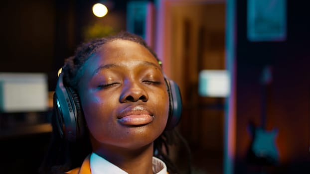 College student sitting laid back after working on class project, relaxing with ambience music on headphones at home desk. African american girl chilling with modern tunes on headset. Camera B.