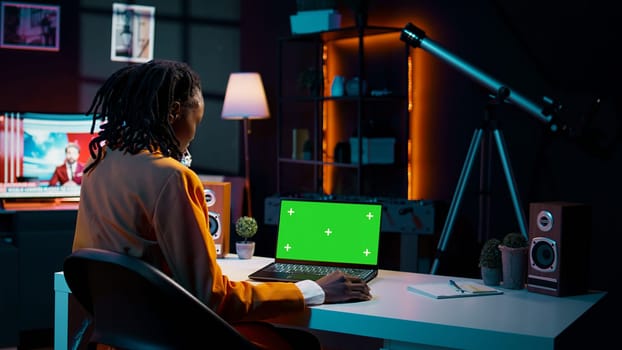 African american young woman looks at laptop with greenscreen, sitting at her home desk and preparing to attend online university lessons. Girl using pc with isolated mockup layout. Camera B.