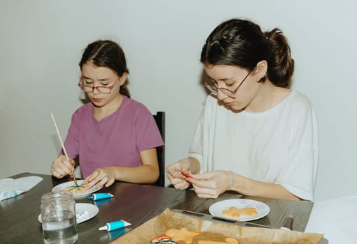Two beautiful Caucasian brunette girls are sitting at the table and enthusiastically decorating baked cookies with paints and chocolate using brushes and tubes, close-up side view. Step by step instructions. Step 25