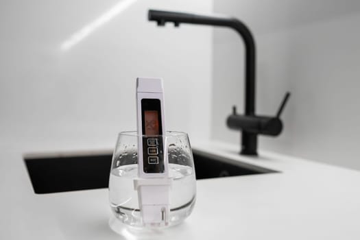 Conductometer or TDS water quality meter is immersed in a beaker of water to check purity in front of modern kitchen water tap. Dirthy water concept